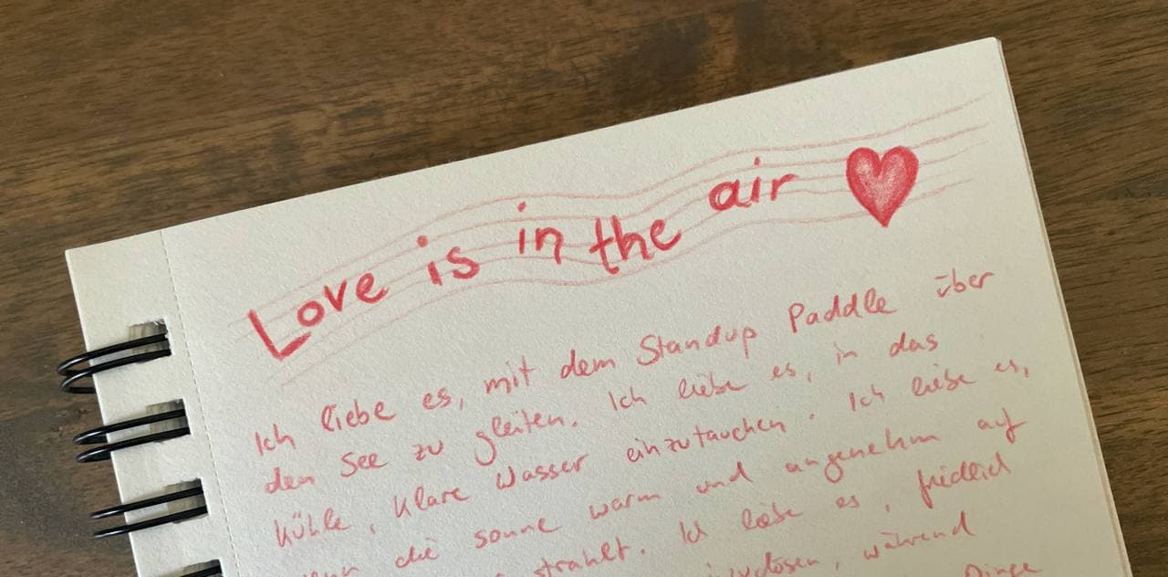 A photo of a sketch book page titled 'Love is in the air' with a few lines of gratitude journaling in German, starting with 'Ich liebe es,...' ('I love it, when...')