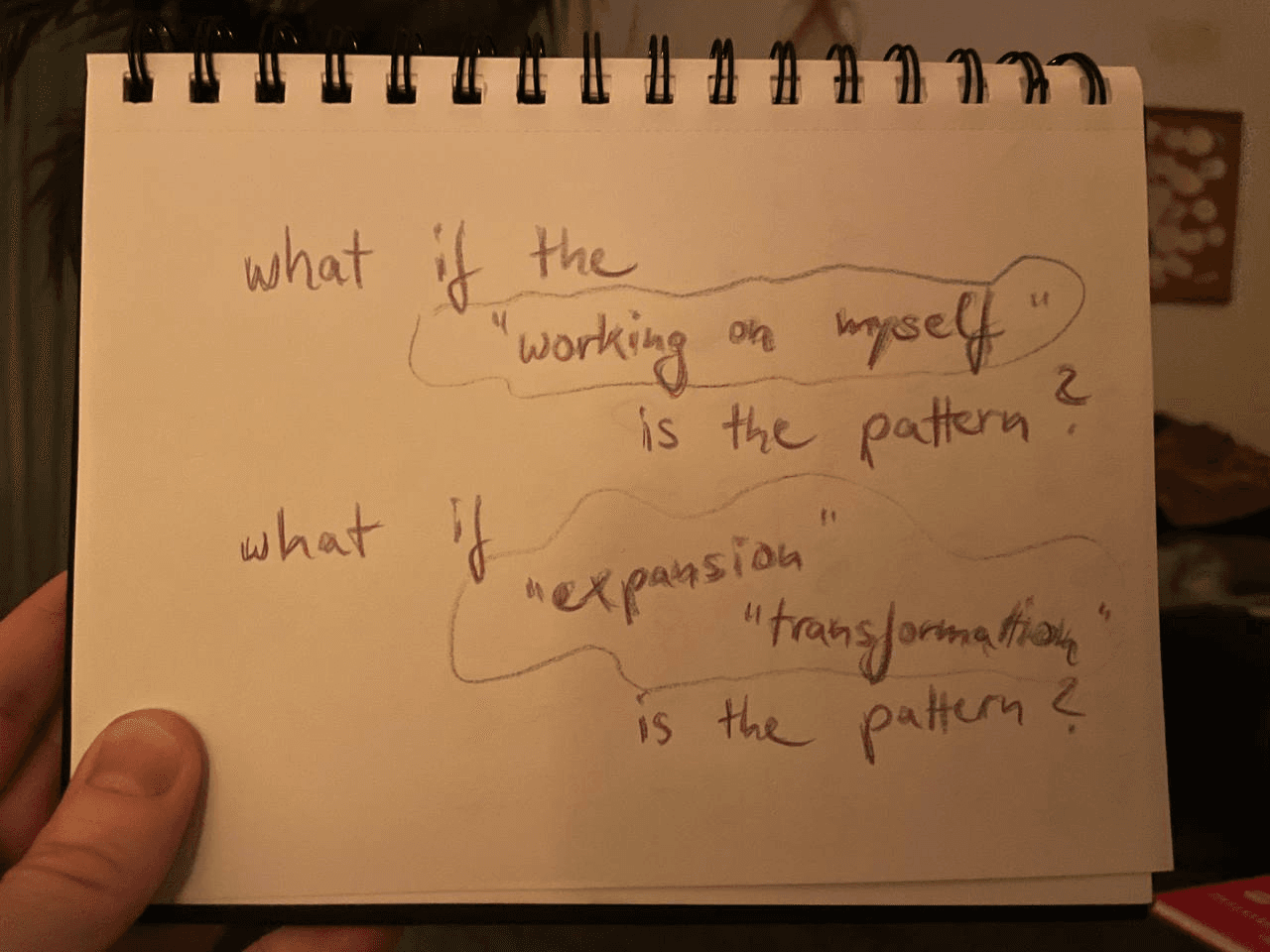 A hand holding a drawing that shows written text: What if the 'working on myself' is the pattern? What if the 'transformation', 'expansion' is the pattern?