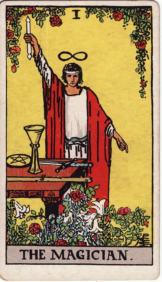 A figure in a red robe stands before an altar with the four suits of tarot laid out before them. They hold a wand pointing upward while pointing downward with their other hand.